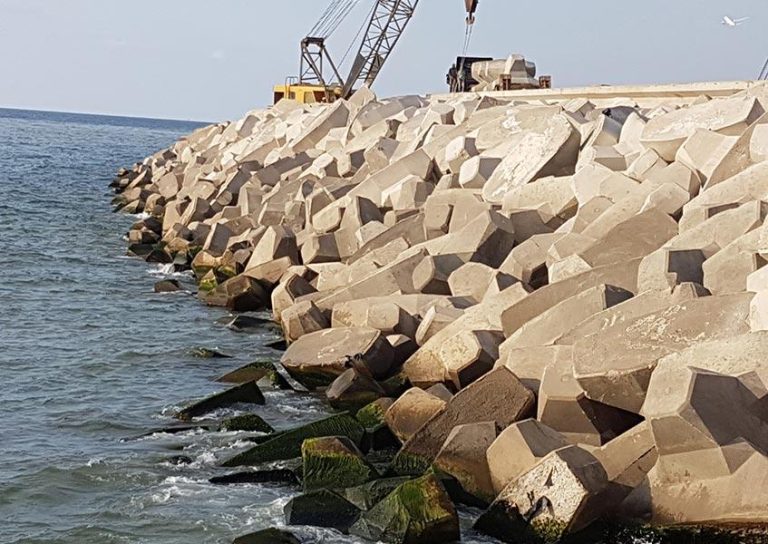 The image shows a coastal area with a breakwater made of large, interlocking concrete blocks. A crane is visible on top of the structure, suggesting construction or maintenance work. The ocean waves lap against the blocks, and some patches of green algae are noticeable.