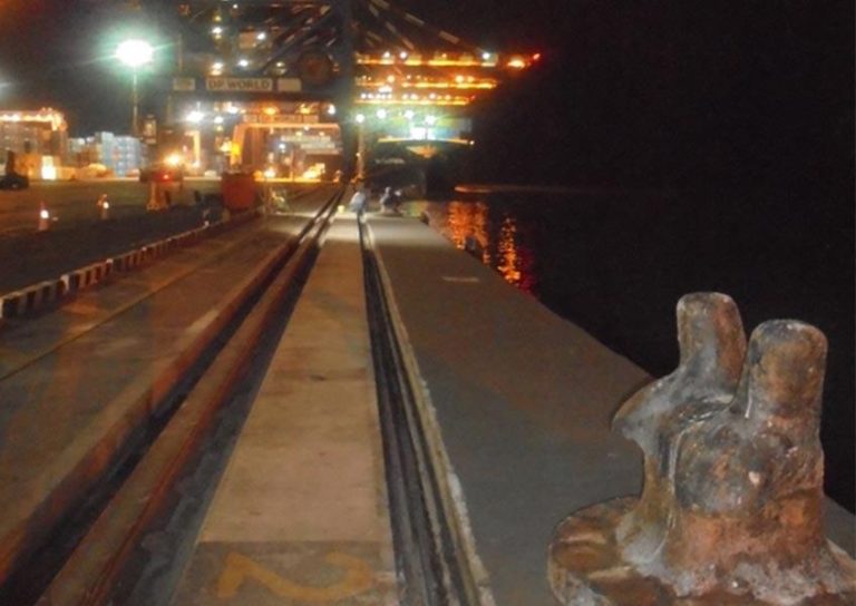 A nighttime photo shows a well-lit port with cranes and stacked shipping containers in the background. In the foreground, there is a dock with metal bollards. The water is dark, reflecting some of the lights from the port.