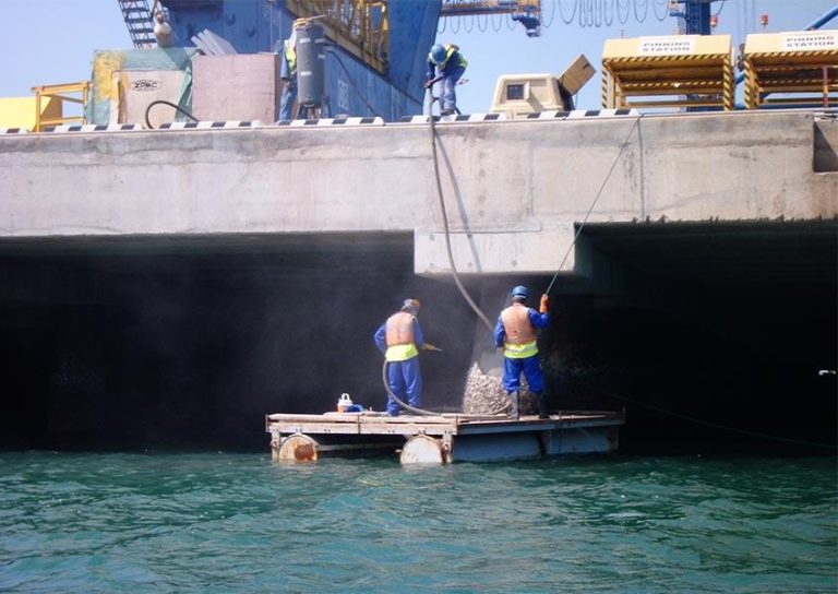 Three construction workers in safety gear work on a concrete structure by the water. Two workers on a floating platform apply material to the underside of the concrete, while the third stands above, holding a hose. The water below reflects the platform.