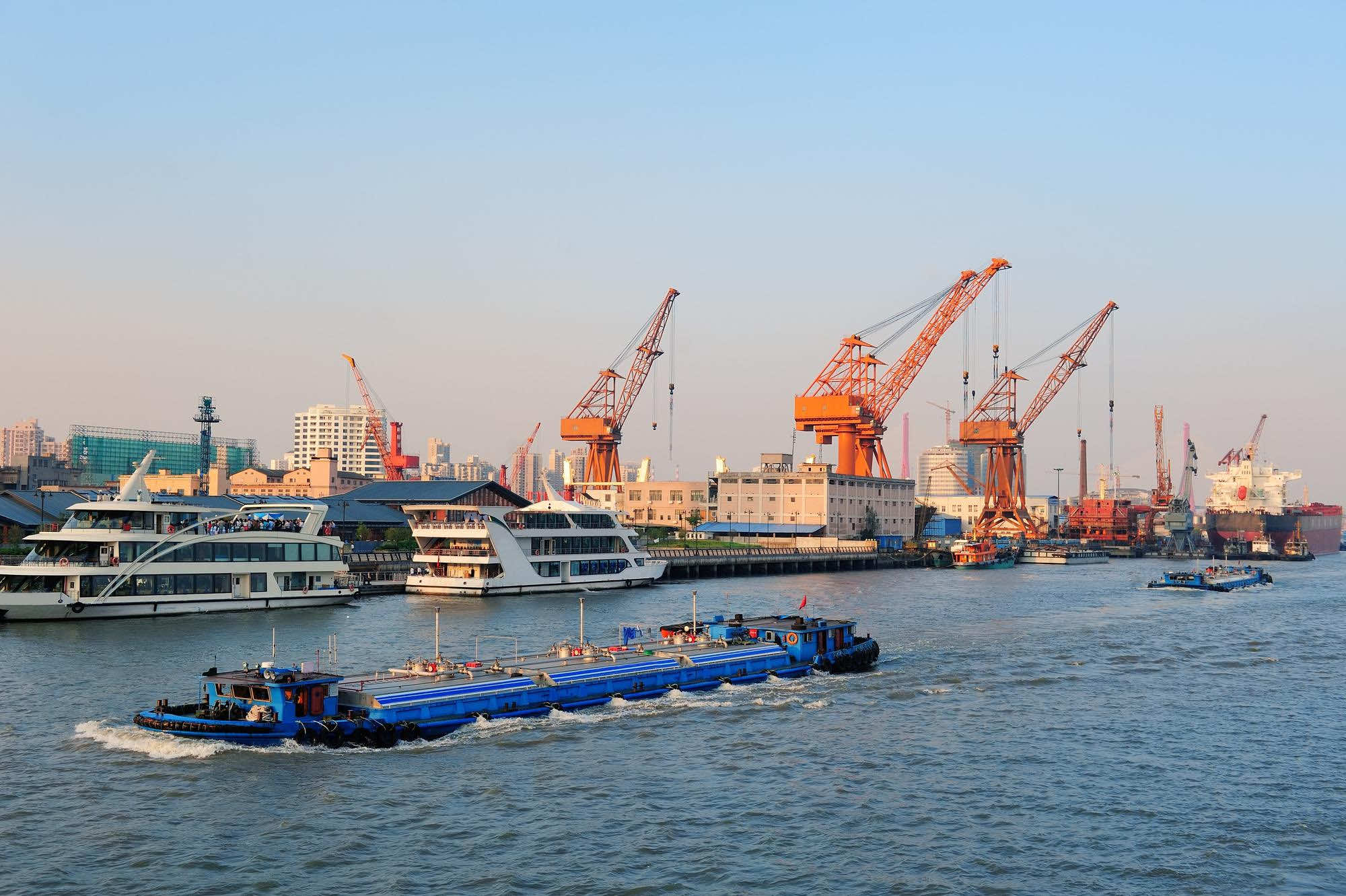 A bustling harbor scene with several cargo ships and barges on the water. Large orange cranes line the dock and buildings are visible in the background under a clear blue sky.