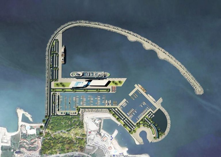 Aerial view of a harbor with a curved breakwater enclosing a marina. The marina has several docks for boats, parking lots, and green spaces. Surrounding the harbor are roads, trees, and buildings. A large vessel is docked in the harbor.