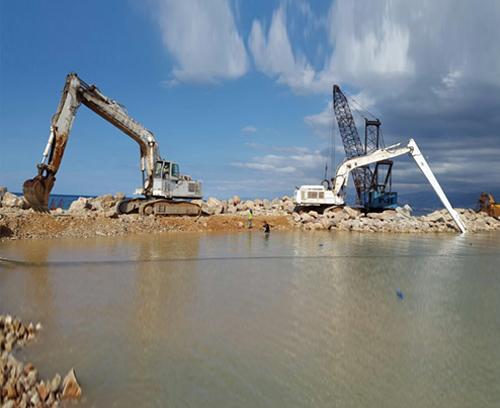 Two large excavators work on a rocky shoreline, digging and moving rocks. One excavator has a long arm extending over water, while the other is closer to the shore, both under a partly cloudy sky. Water and rocks dominate the foreground.