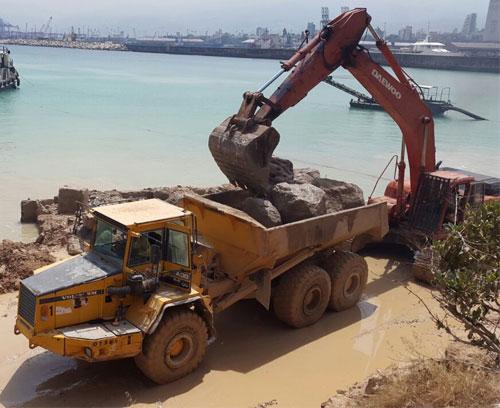 An orange excavator with "Doosan" written on its arm is loading large rocks into a yellow dump truck. The vehicles are on a sandy shoreline, with a body of water and industrial structures visible in the background.