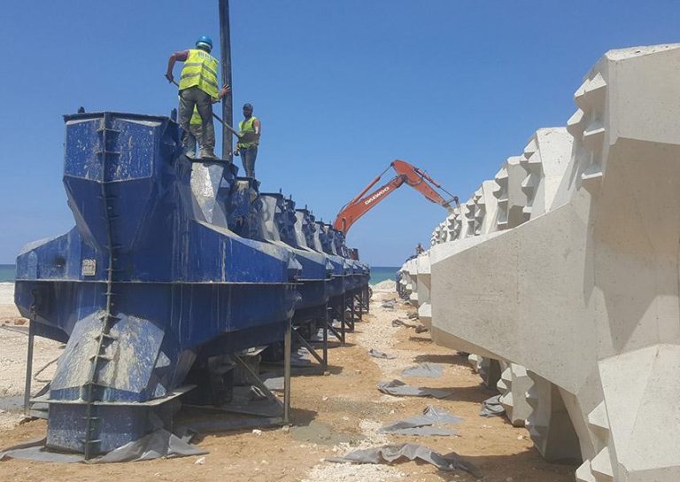 Construction workers in safety vests and helmets are standing on large blue metal structures. A heavy-duty excavator operates in the background, adjacent to rows of angular, white concrete components. The scene takes place under a clear blue sky.