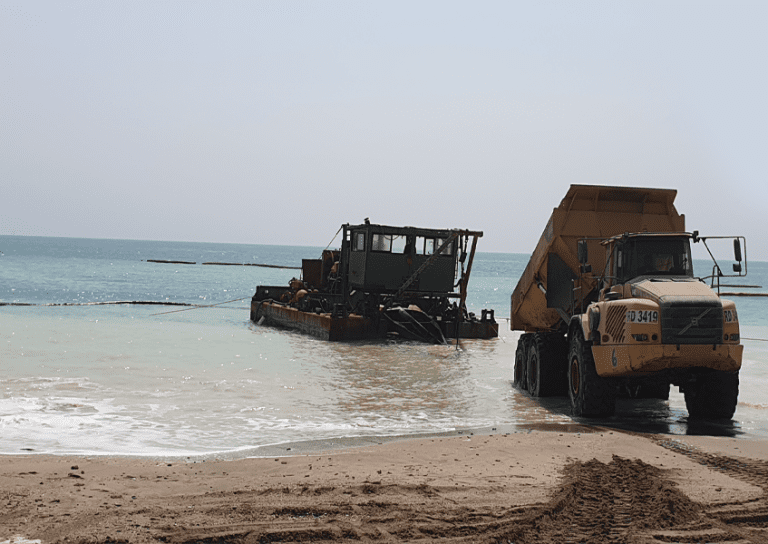 A barge is stationed in shallow turquoise waters near a beach, connected by a series of hoses to a large yellow dump truck on the sandy shore. The clear sky and calm sea create a serene backdrop for this scene of marine construction or dredging work.