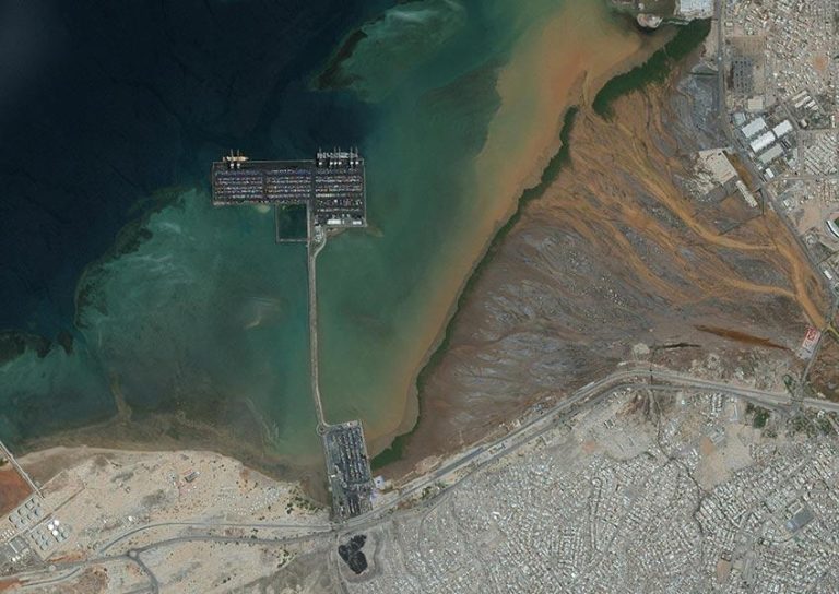 A satellite image showing a coastal area with a large dock extending into a dark blue body of water. Brown, murky river plumes mix with greener waters near the shore. Beneath, urban and industrial areas with roads and buildings are visible in the lower part of the image.