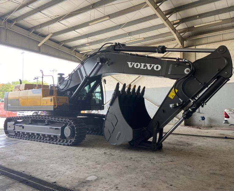 A large Volvo excavator with a digging bucket attachment is parked inside a spacious warehouse-like structure. The machine is black and yellow, with visible tracks for movement. The surrounding area includes concrete flooring and overhead metal beams.