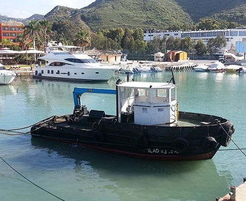 A small, black tugboat with a blue crane is docked at a marina. In the background, there are several white yachts, palm trees, and buildings along the waterfront, with a backdrop of green, hilly terrain.