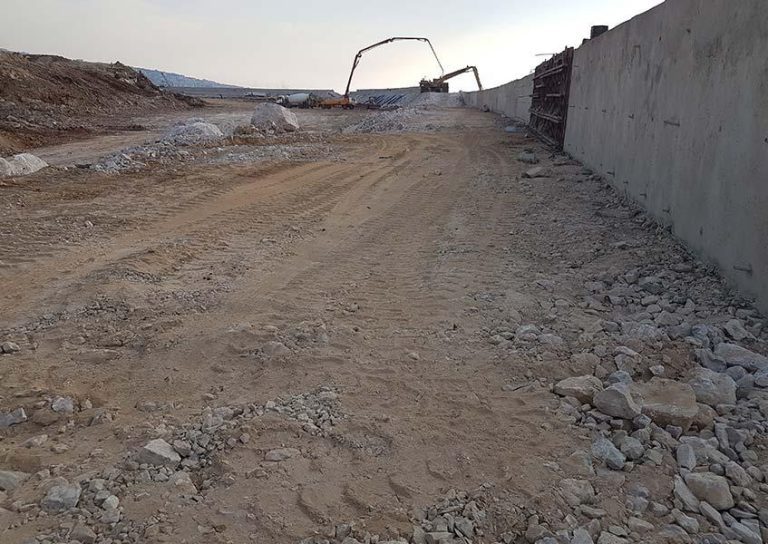 A construction site showing a partially built road with a concrete wall on the right side. Heavy machinery is visible in the distance, and the ground is covered with loose dirt and rocks. The sky is clear with a slight haze.