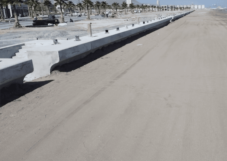 A wide concrete promenade under construction along a sandy beach with rows of newly planted palm trees on the left. The sky is clear, and there is some construction equipment in the background.