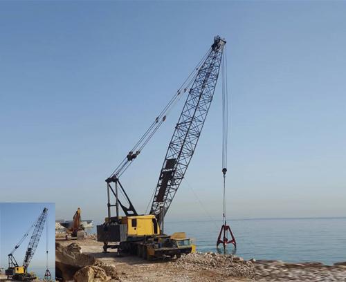A yellow construction crane situated on a coastal area with a rocky terrain, lifting materials with a jib-arm extended over the calm, blue sea. Clear blue sky in the background. A small inset image displays the same crane from a slightly different angle.