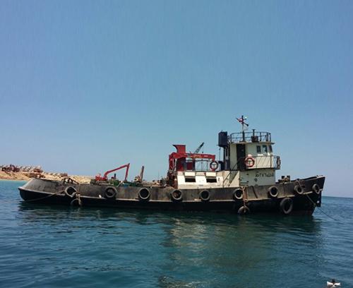 A weathered tugboat floats on calm, clear blue water under a bright sky. The boat has a red crane on its deck and various equipment. In the background, there is a rocky shoreline.
