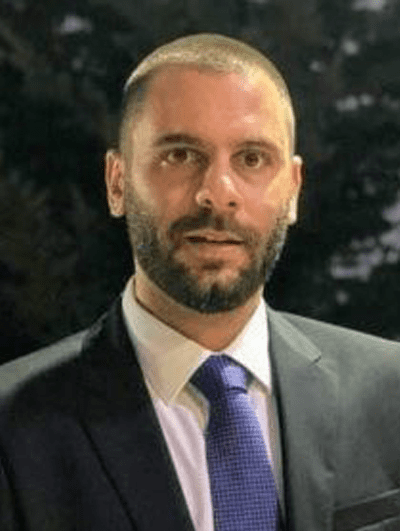 A man with a closely cropped beard and short hair, wearing a dark suit, white shirt, and blue tie. He is looking ahead with a neutral expression. The background is blurred and appears to be outdoors at night.