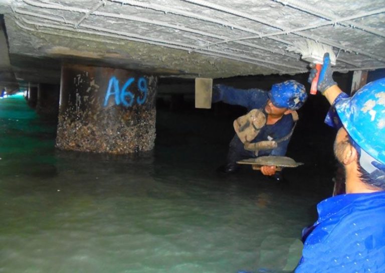Two workers wearing blue hard hats and safety gear are repairing the underside of a concrete structure above water. One is applying a substance with a brush, while the other holds a trowel. Water surrounds their legs; a column labeled "A69" is visible in the background.