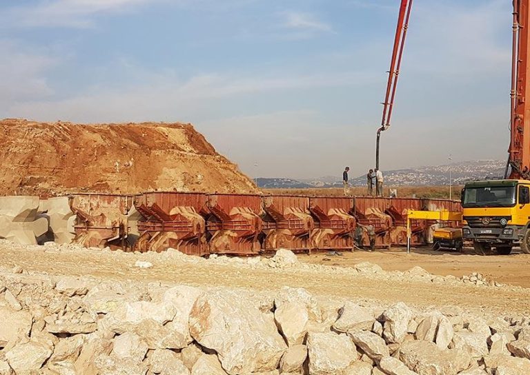 A construction site with large machinery and workers. A rusted metal structure is in the foreground, and a truck is parked nearby. The background features a large mound of dirt and rocks, with scattered rocks in the foreground under a partly cloudy sky.