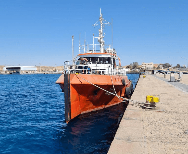 A bright orange boat is tethered to a dock on a sunny day. The dock stretches along the right side of the image, while the deep blue water and a clear, cloudless sky fill the background. Buildings and structures are visible in the distance.