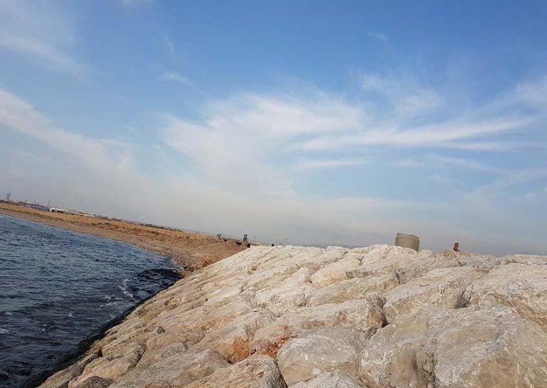 A rocky shoreline stretches out under a blue sky with scattered clouds. The rocks form a barrier between the water and the land, which extends into the distance. The sea is calm, and the coastline curves slightly.