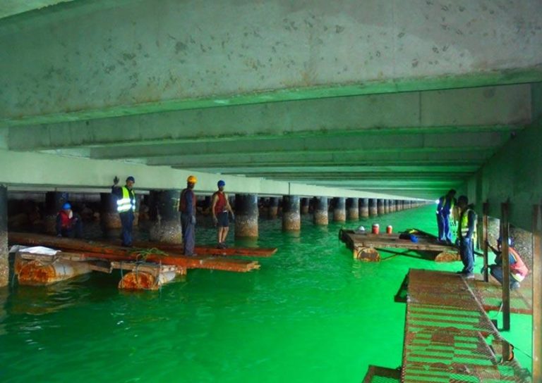 Several workers are standing on platforms under a large bridge with multiple pillars emerging from green water. Some workers wear safety vests and helmets, indicating ongoing construction or maintenance activities. The image depicts an industrial and aquatic environment.