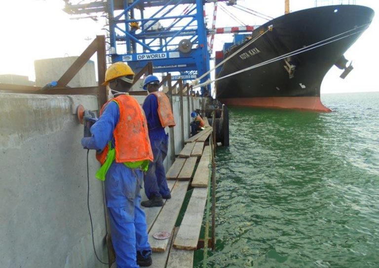 Construction workers in orange vests and yellow helmets work on a concrete structure at a dock, using power tools near the water. A large cargo ship is docked nearby under a blue crane labeled "DP World." The scene overlooks a body of water.