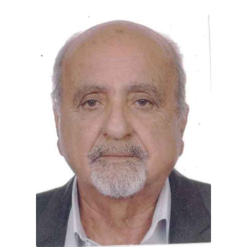 A headshot of an older man with a bald head, gray hair on the sides, and a mustache and beard. He is wearing a white shirt and a dark-colored jacket, looking straight at the camera with a neutral expression. The background is plain white.