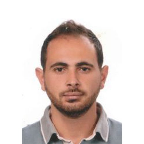 A man with short dark hair and a trimmed beard is looking directly at the camera. He is wearing a grey collared shirt with a light grey trim. The background is plain white.
