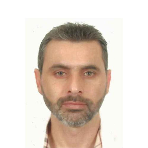Portrait of a man with short, graying hair and a beard, against a plain white background. He has a serious expression and wears a checkered shirt.