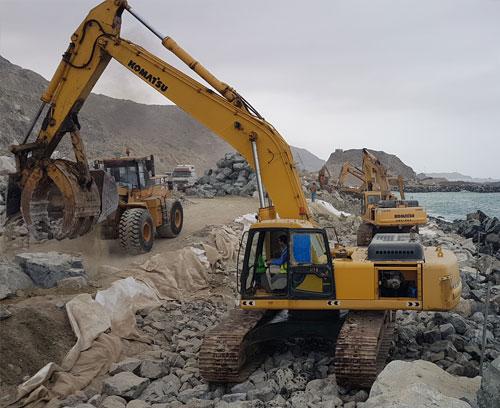 Three excavators are working on rocky terrain near a body of water. The construction equipment are yellow, and the workers are maneuvering them to move large rocks. In the background, there are steep hills and more construction activity taking place.
