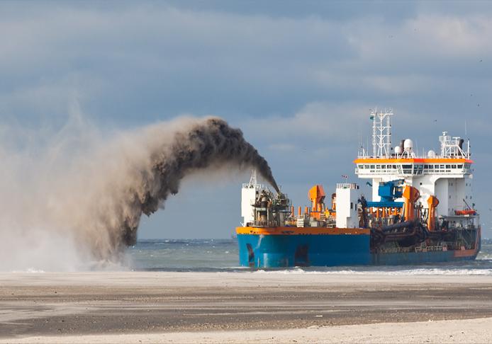 A large blue and orange dredging ship operates near a sandy beach, expelling a large plume of dark sediment back into the sea. The sky is cloudy, and the choppy waters illustrate the ship's ongoing dredging activity.