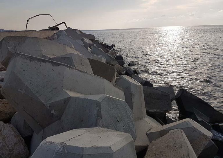 A coastal breakwater made of large, irregularly shaped concrete blocks stretches into the ocean. The sun is low, casting a bright reflection on the water. A crane is visible in the distance, suggesting ongoing construction or maintenance.