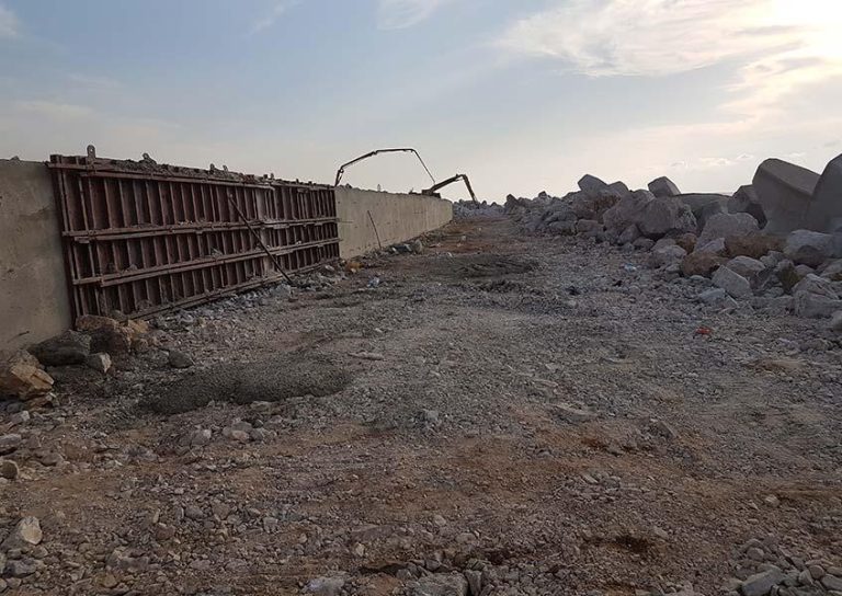 A construction site with a partially built concrete wall on the left, supported by wooden frames. The ground is covered with gravel and rocks, and large boulders are scattered on the right. A cloudy sky can be seen in the background.