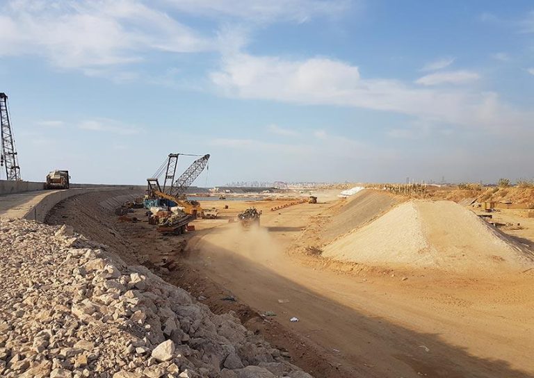 A construction site with cranes, excavators, and a dump truck. Dust is being kicked up by machinery working on uneven terrain. The sky is clear with scattered clouds, and there are piles of dirt and gravel beside the main work area.
