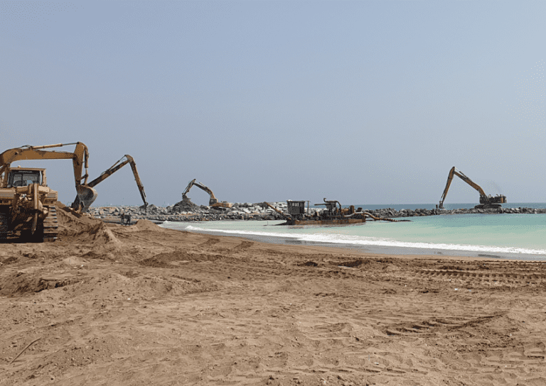 A beach construction site with several excavators and heavy machinery working on a rocky breakwater. The machinery is positioned near the shoreline, and piles of rocks are being moved. The sea is visible in the background under a clear, blue sky.