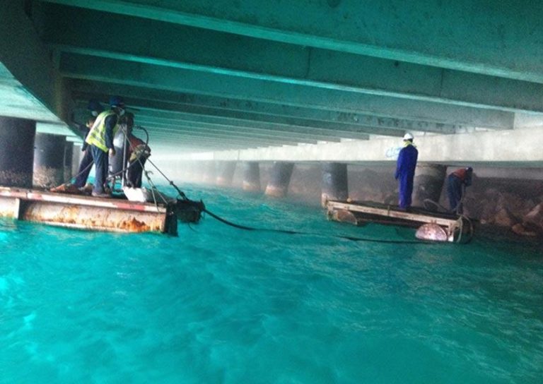 Workers in safety gear operate equipment underneath a large bridge structure. One worker is on a metal platform on the left, and two others are on another floating platform on the right. The water below is a vibrant turquoise color, and the area is misty or foggy.