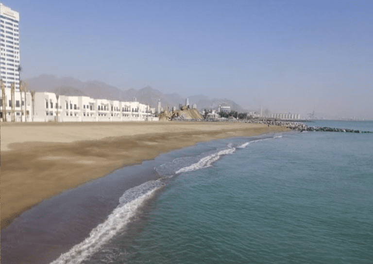 A serene beach scene with gentle waves lapping the shore. White buildings line the coast with tall mountains in the background. Off in the distance, a breakwater of rocks extends into the calm, blue sea under a clear sky.