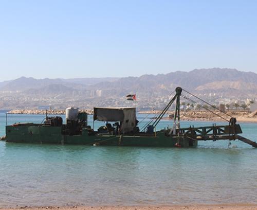 A dredging vessel with a crane is positioned in shallow water near the shore. The vessel has a canopy and equipment onboard. Background includes a clear blue sky, distant mountains, and a shoreline with buildings.