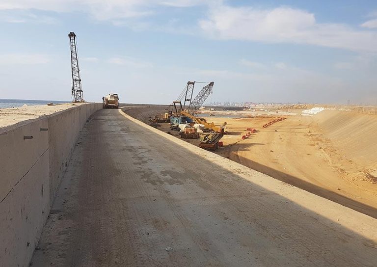 A construction site featuring heavy machinery, including cranes and bulldozers, working along a dirt-filled area next to a concrete pathway. The sky is partly cloudy, and the distant cityscape is visible on the horizon.