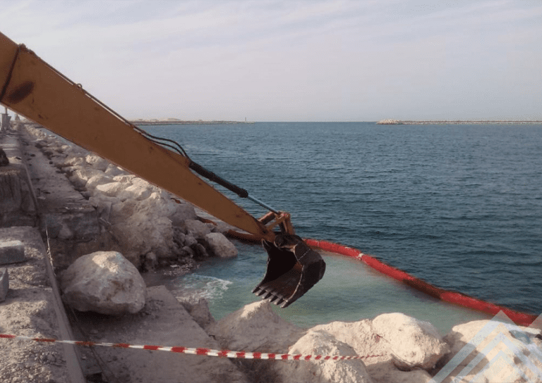 An excavator with its arm extended is positioned near a rocky shoreline. The machine's bucket hovers above the water, and a red floating barrier separates the construction area from the sea. The sky is partly cloudy, and the water appears calm.