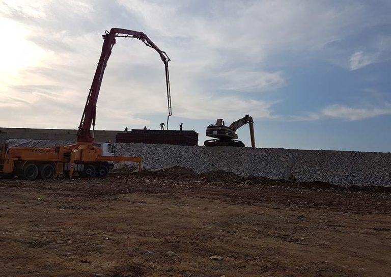 A construction site with a crane lifting materials and a bulldozer moving on a gravel-covered area. Workers are visible on a structure in the background against a partly cloudy sky. The ground is a mix of dirt and gravel.