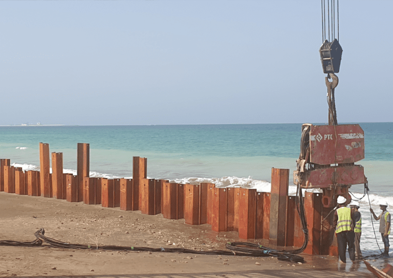 Construction workers install sheet piles with heavy machinery along a coastline. Several large, rust-colored sheet piles are already positioned in the sand near the water's edge, with waves gently crashing. The ocean and a clear sky form the background.