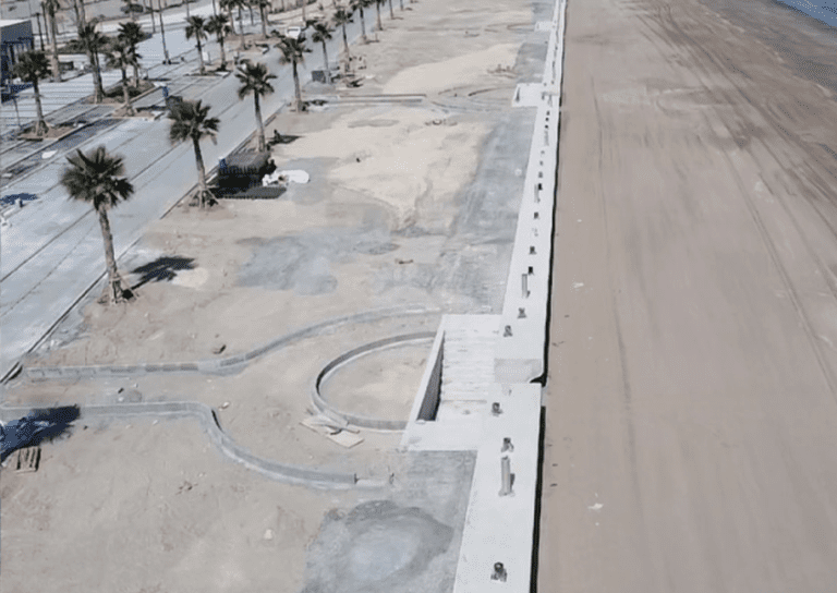 Aerial view of a beachfront promenade under construction. Palm trees line the pathway on the left, and there are unfinished concrete structures along the beach. The right side shows a wide expanse of sandy shoreline, while the ocean is out of frame.