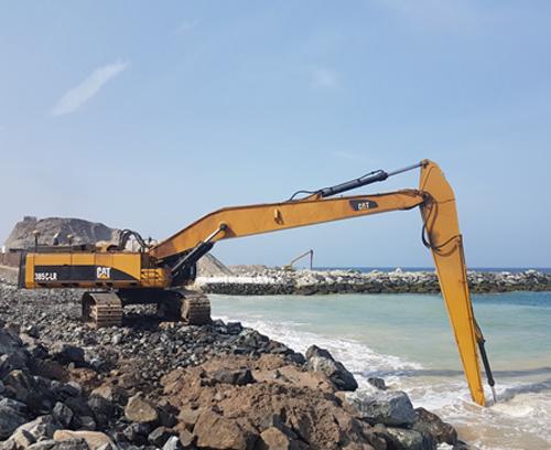 A long-reach Caterpillar excavator with yellow and black paint sits on a rocky shoreline, extending its arm and bucket towards the water. The backdrop includes a clear blue sky, an ocean, and a distant rocky structure.