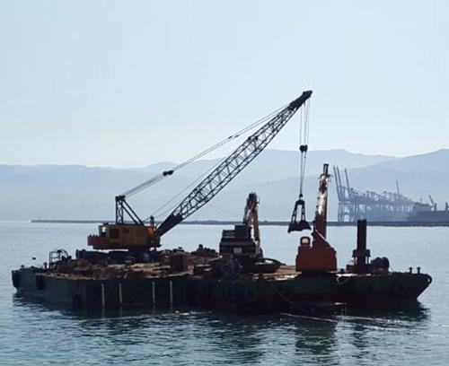 A barge equipped with a large crane is floating on calm waters with a coastline and mountains in the background. The crane is extended over the side of the barge, lifting a piece of equipment. Industrial structures are visible in the distance.