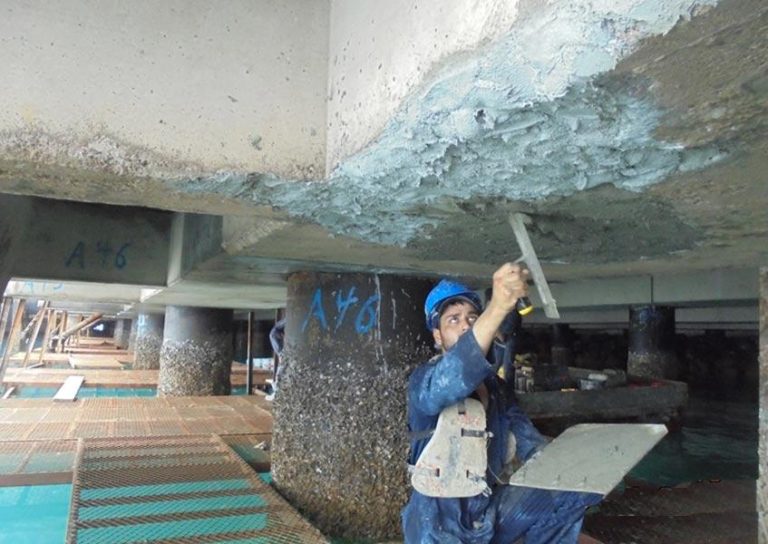 A construction worker wearing a blue hard hat and safety vest is repairing a concrete structure under a bridge. They are smoothing a gray material onto the surface using a trowel. The worker is standing on a platform above water, surrounded by concrete columns.