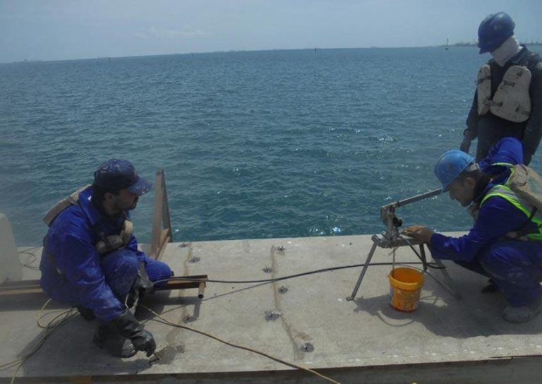 Three workers in protective gear are conducting maintenance or inspection on a concrete platform by the sea. One operates a piece of equipment attached to the platform, another holds a tool, and a third observes. An orange bucket is placed next to the equipment.