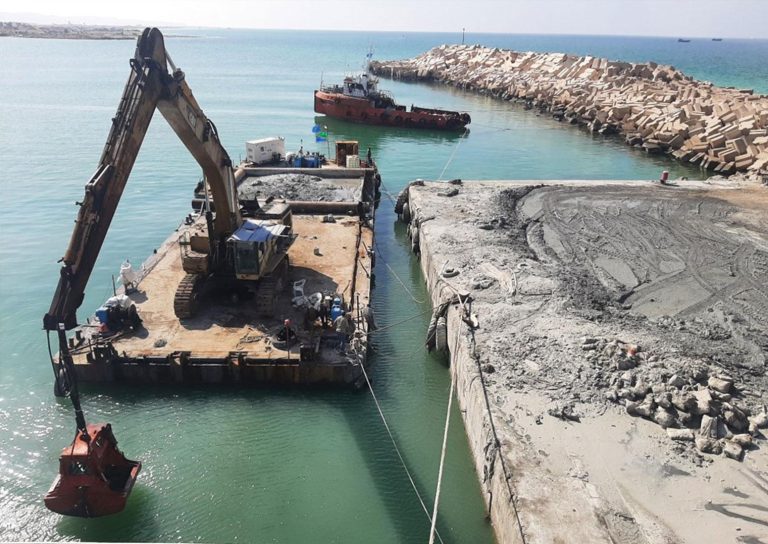 An excavator on a barge dredges a harbor, with a tugboat and breakwater in the background. Construction materials and equipment are visible, and another section of the harbor shows piles of rubble. The water is a clear blue under a sunny sky.