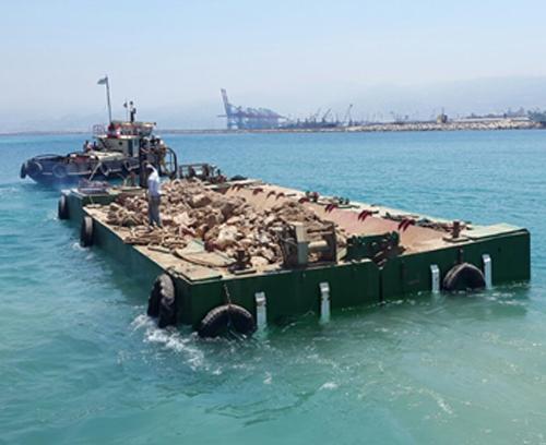 A barge carrying a load of rocks sails through a calm, turquoise sea. A person stands on the barge, and in the background, a port with cranes and industrial structures is visible under a clear sky. The boat appears to be guided by a tugboat.