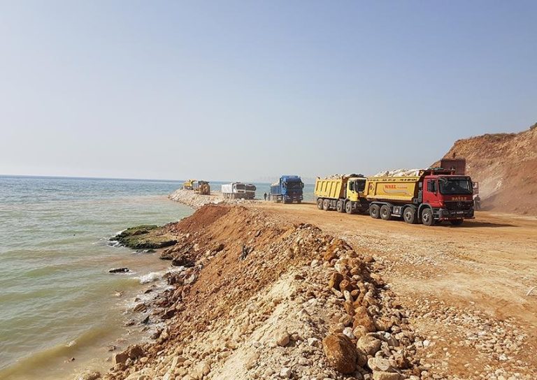 Several large construction trucks are parked along a dirt road next to a body of water. The trucks appear to be involved in a coastal construction or excavation project. The scene includes rocky terrain, piles of soil, and a clear blue sky.