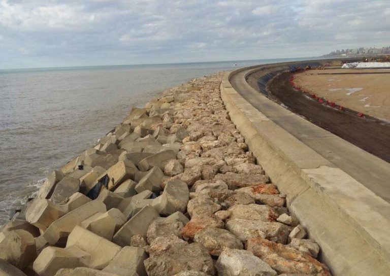 A long seawall made of large rocks and concrete barriers curves along the shoreline under a partly cloudy sky. Waves crash lightly against the structure, and a dirt path runs parallel to it on the right, bordered by red safety barriers.