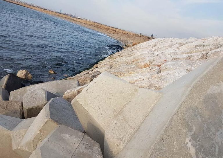 A coastal scene with large concrete blocks forming a breakwater extending into the sea. The shoreline is lined with smaller rocks and sand, and the sky is slightly overcast. A person is barely visible in the distance near the tip of the breakwater.