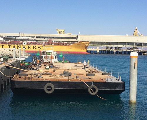 A flat-deck barge is docked at a pier in a harbor. In the background, there are industrial buildings and a partially visible large tanker ship with the label "TANKERS." Workers and equipment are present on the barge, and the sky is clear and blue.